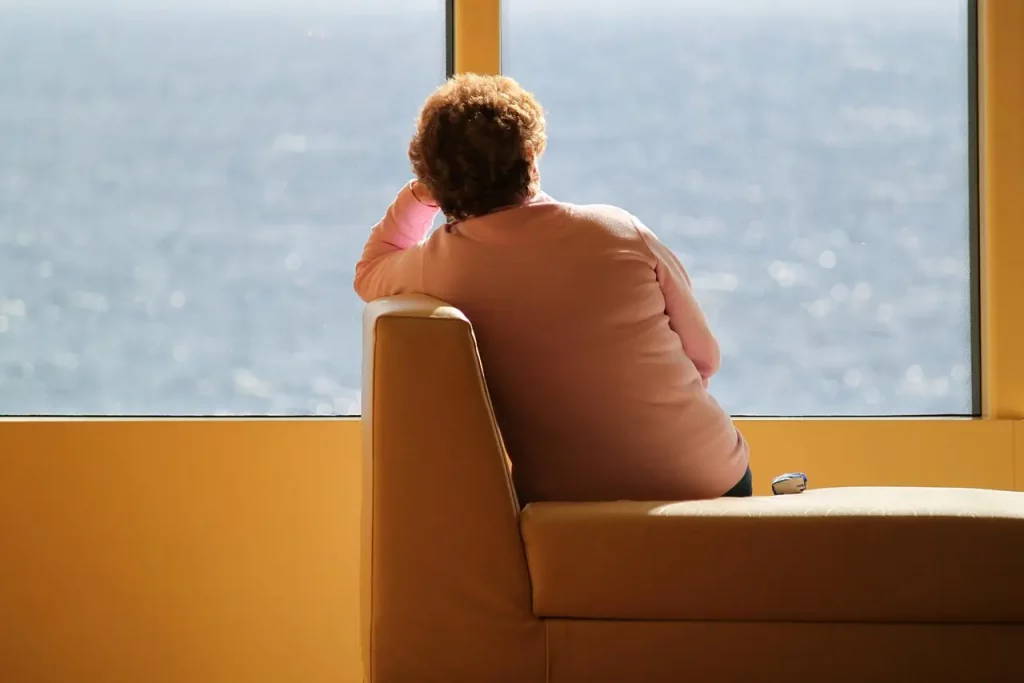 Une femme d'un certain âge, vue de dos, est assise seule sur une banquette. Son regard est perdu à travers la fenêtre, fixant l'horizon de la mer lointaine. Dans cette posture solitaire, elle semble absorbée par ses pensées, laissant transparaître un profond sentiment de solitude qui pèse sur elle comme une vague implacable.
