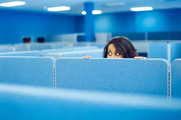 Cette image captivante nous montre une scène dans un environnement de bureau moderne. Une femme est assise à son bureau dans un open space, mais seule sa tête dépasse de la cloison. Son expression faciale révèle une profonde inquiétude alors qu'elle regarde nerveusement à droite et à gauche, comme si elle redoutait l'arrivée de quelqu'un. Cette illustration met en lumière les tensions et les craintes souvent ressenties dans les milieux professionnels, où la peur de l'évaluation, du jugement ou des conséquences peut être omniprésente. Comprendre les origines et les besoins liés à la peur dans les organisations est essentiel pour promouvoir un environnement de travail plus sain et plus productif, où chacun se sent en sécurité et soutenu.