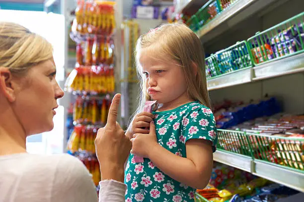 Dans cette scène saisissante, une mère se tient dans un magasin, son enfant à ses côtés. Son visage exprime la frustration alors qu'elle réprimande sa fille, pointant un doigt avec fermeté. Cette image évoque les défis de la parentalité et les messages contraignants transmis de génération en génération, soulignant l'importance de reconnaître et de briser ces schémas pour cultiver une estime de soi saine.